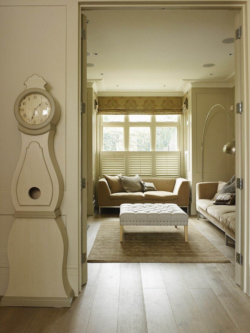 Swedish longcase clock next to open door with view of sofa set below window in rustic interior with pale colour scheme