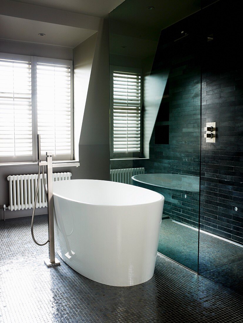 weiße, freistehende Designer Badewanne mit Standarmatur auf dunklem Mosaikboden und verglaster Duschbereich