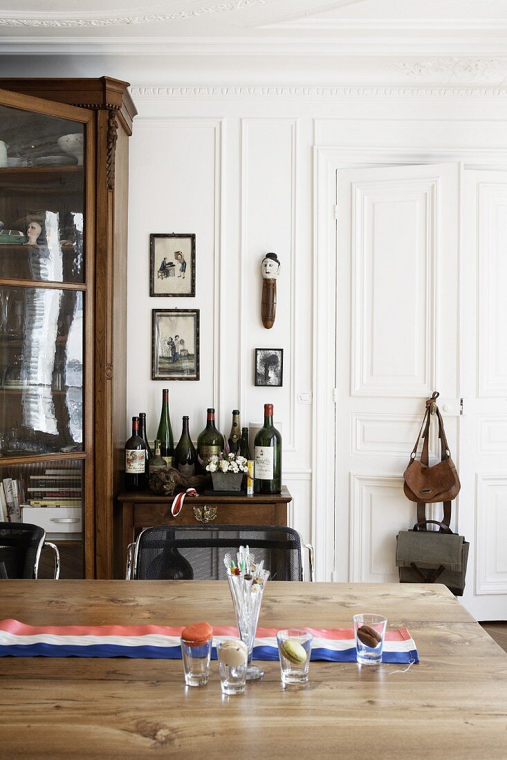 Holztisch mit französischem Fahnenband, im Hintergrund Beistelltisch mit Weinflaschen und antiker Vitrinenschrank
