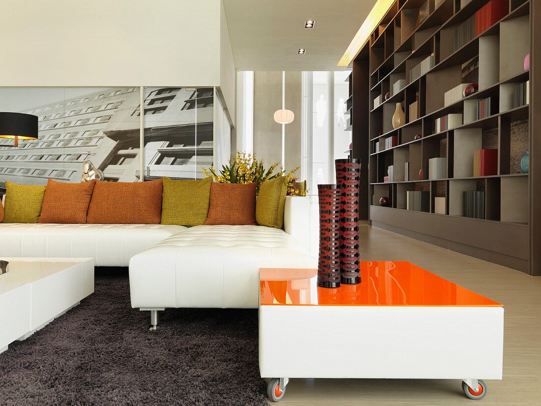 Ein modernes Wohnzimmer mit weißem Ecksofa und rollbarem Beistelltisch mit orangefarbener Oberfläche vor einer urbanen Fotografie an der Wand
