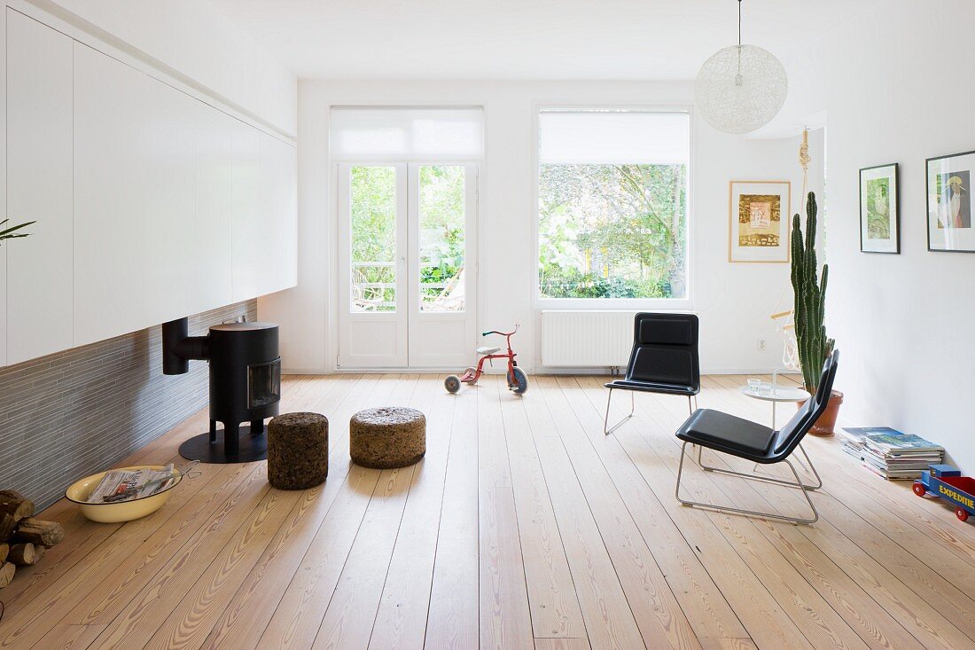 Minimalistisches Wohnzimmer mit schwarzen Sesseln auf Dielenboden und Sitzpolster um kleinen Kanonenofen vor Terrassentür mit Gartenblick
