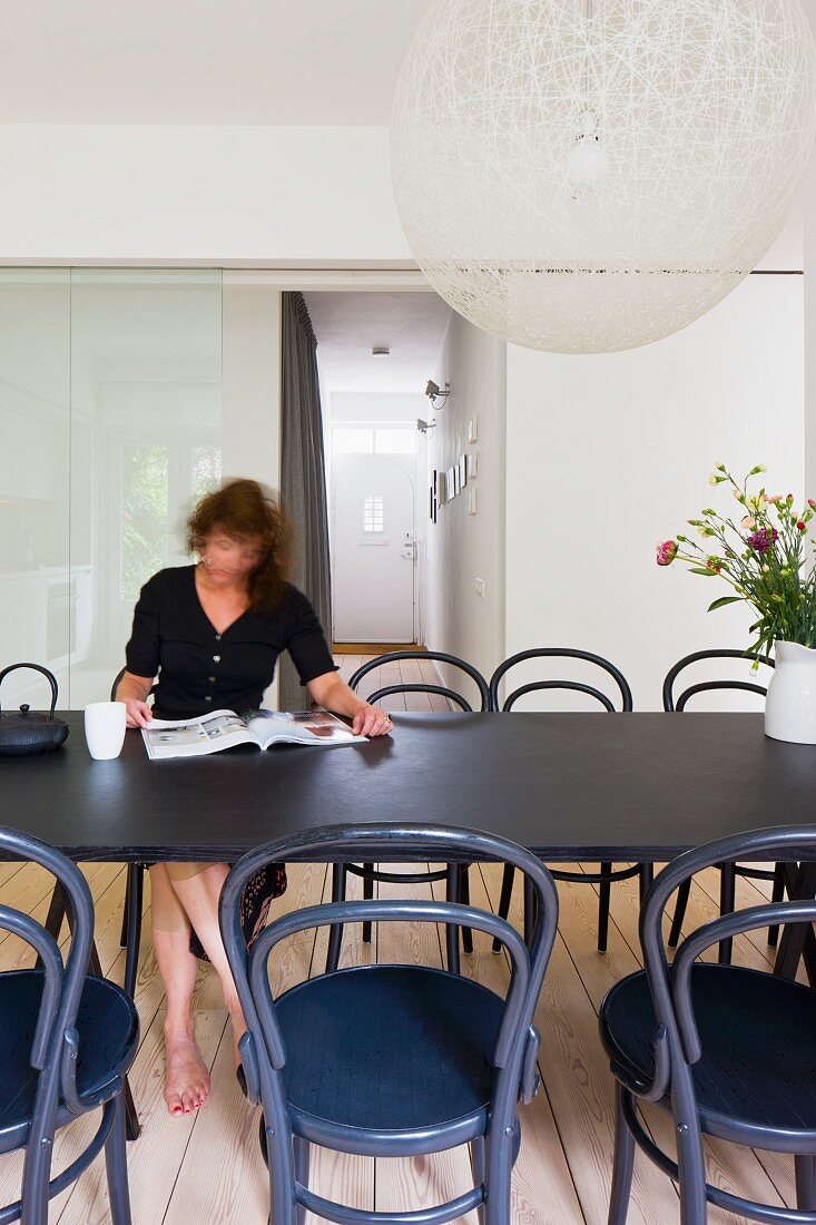 Frau liest am Esstisch - Kugelförmige Designer Hängeleuchte aus weißem Geflecht über Tisch mit schwarzen Thonetstühlen