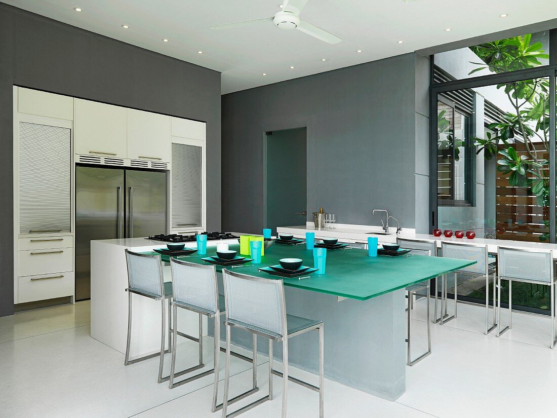 Minimalistische Designerküche mit Kochinsel und Barhockern am gedeckten Glastisch