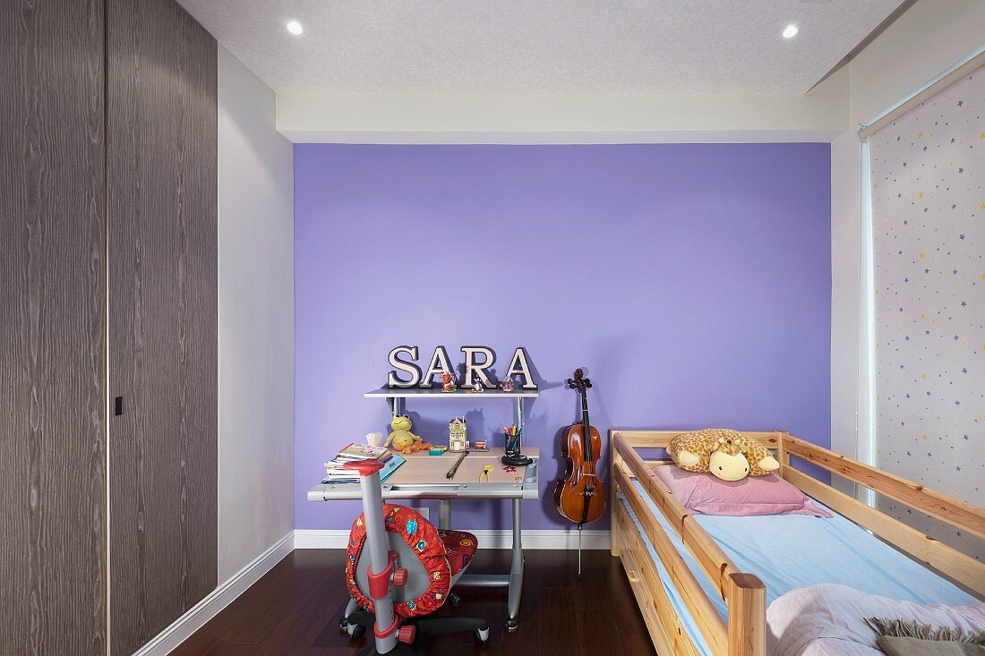Bett vor einem Fenster mit Stoffrollo und Schreibtisch vor lavendelblauer Rückwand in schmalem Kinderzimmer