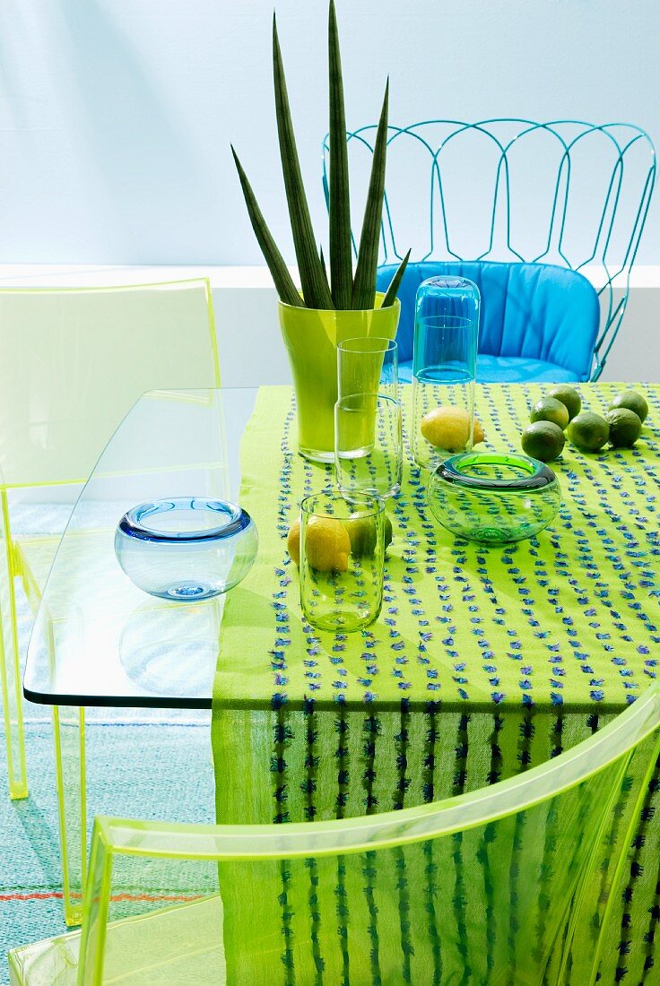 Glastisch mit aquafarbener Tischdeko, Drahtstuhl mit blauem Stitzpolster & grünen Plexiglasstühlen