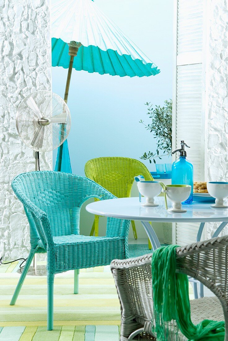 Set of wicker chairs in turquoise, green and white at round table; fan and parasol in front of sky blue wall in background