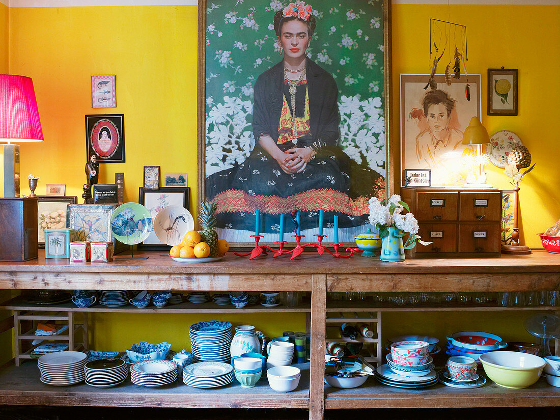 Grosses Frida-Kahlo-Selbstbildnis zentral über offenem Geschirrregal; umgeben von verschiedenen Bildern und Porzellanmalereien vor gelber Wand