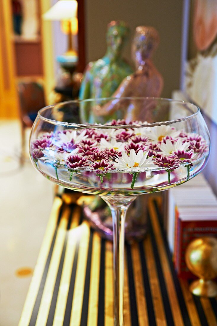 Champagnerschale mit schwimmenden Blüten auf vergoldeter Ablage; im Hintergrund Keramikfiguren