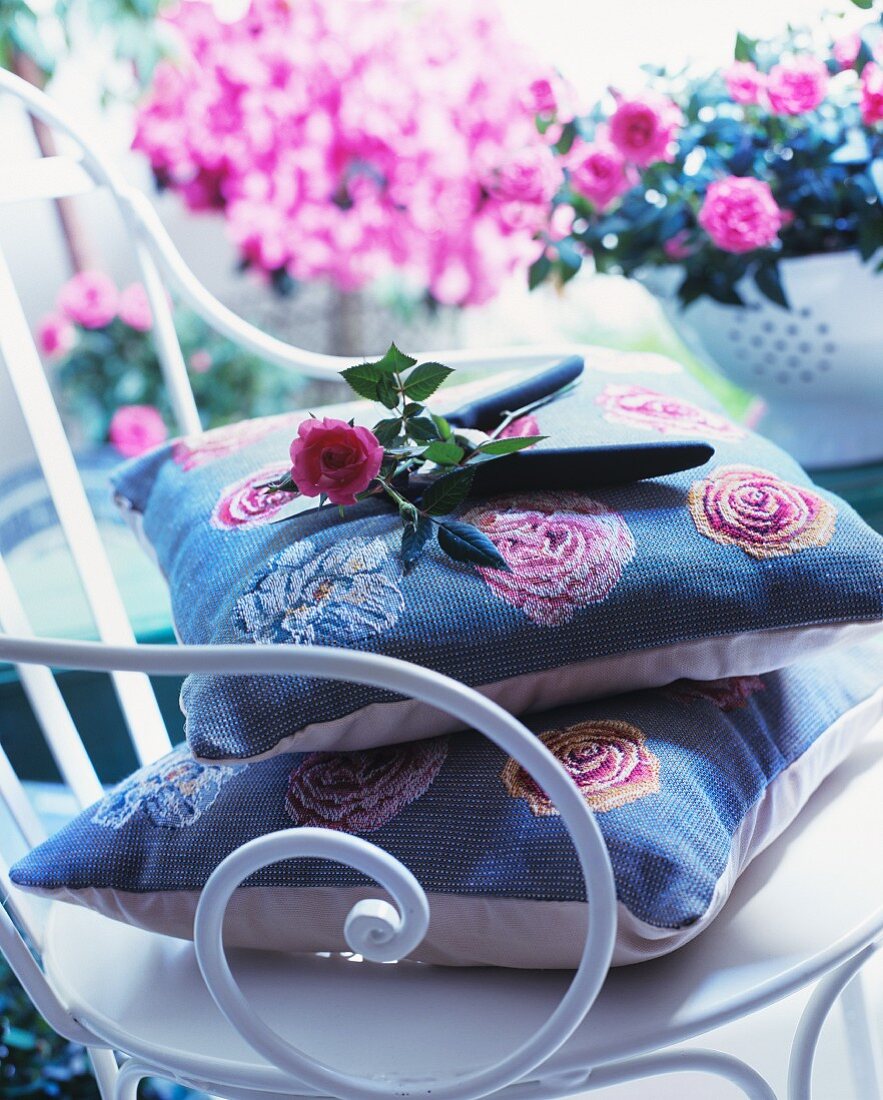 Rosenblüte und Gartenschere auf Kissen und weißem Metallstuhl im Vintagegestil vor Blumentöpfen