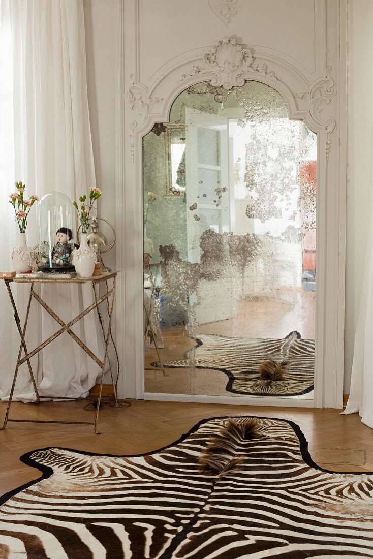 Side table next to antique mirror and zebra-skin rug on parquet floor in bedroom
