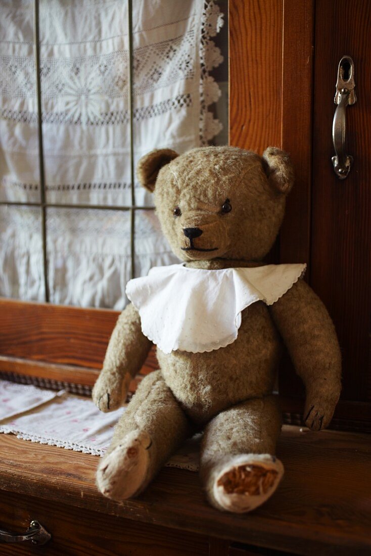 Alter Teddybär mit Lätzchen auf Küchenbuffet vor Spitzenvorhang