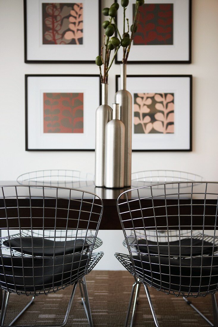 Esstisch mit metallenen Gitterstühlen und glänzenden Blumenvasen; im Hintergrund vier quadratische Wandbilder