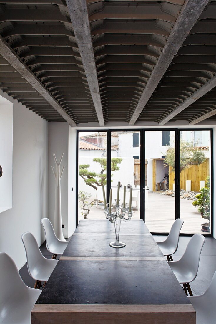 Transparenter Glaskandelaber und weiße Eames Stühle am zeitgenössischen Esstisch eines Wohnraums mit Fensterfront zur Terrasse und auffälliger Stahl-Holz-Deckenkonstruktion