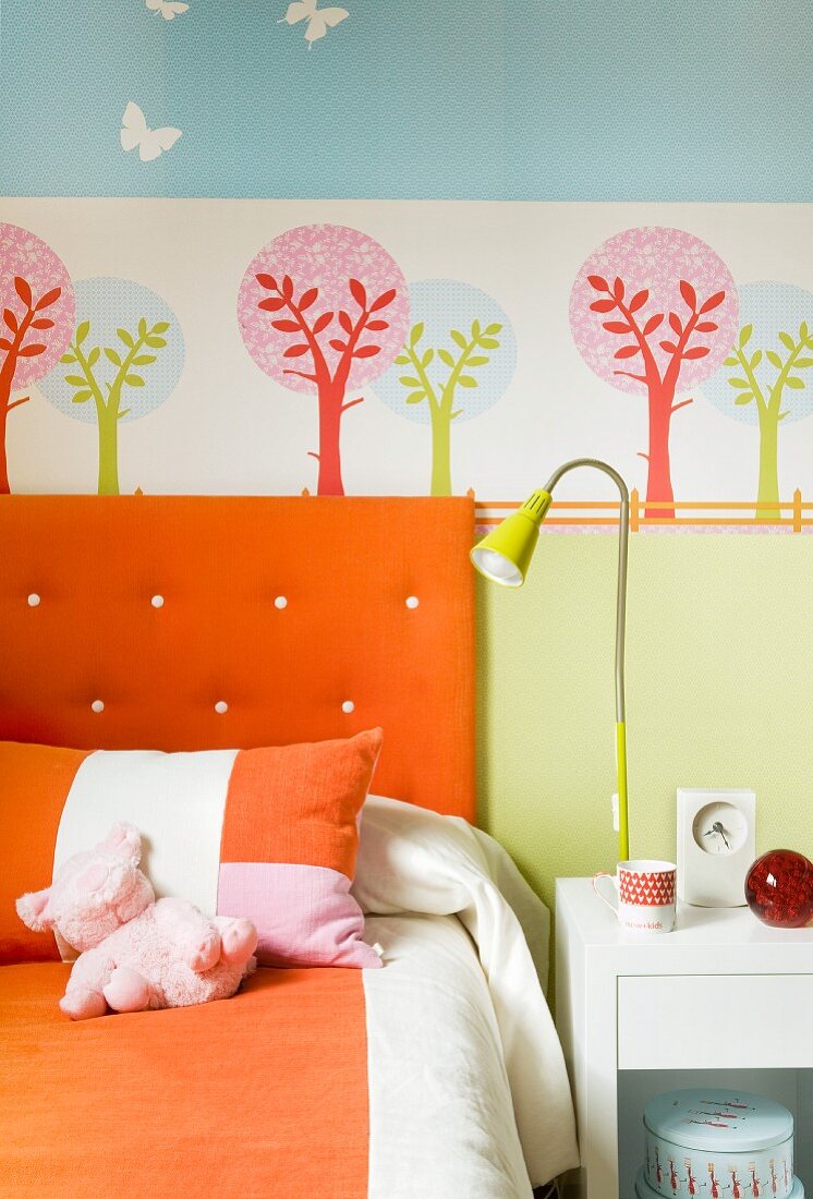 Colorful child's bedroom - orange upholstered headboard and bed linen in front of a tree wall stencil