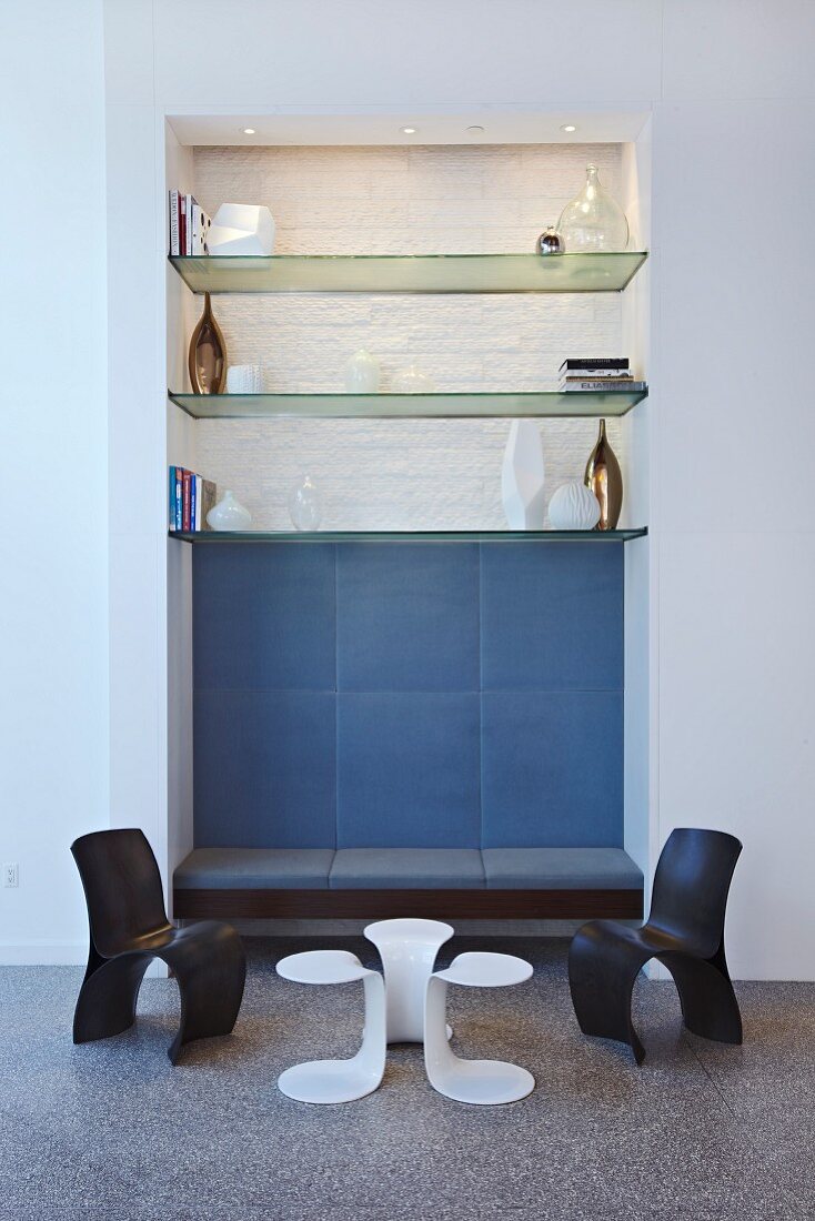 Multifunktionale Wandnische mit Glasregalen und integrierter Polsterbank hinter kleiner Sitzgruppe im Designerstil