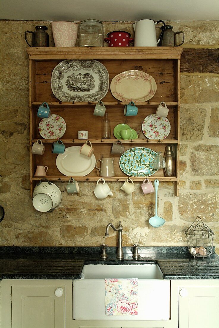 Nostalgisches Küchenregal mit diversen Tellern, Tassen und Kännchen an Haken aufgehängt über dem Vintagespültisch an der Natursteinwand