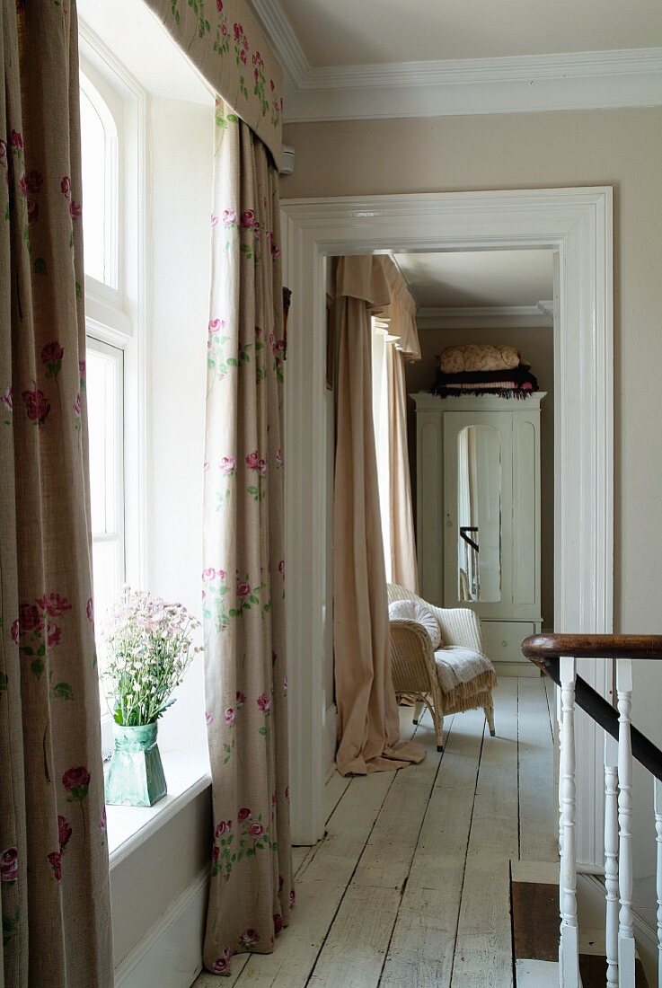 Blick vom Treppenabgang in offenen Raum mit gepolstertem Korbsessel und antikem weißem Kleiderschrank, bodenlange Vorhänge zieren die Fenster und wirken verspielt