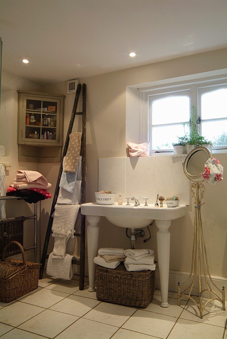 Washstand with legs next to round mirror on delicate metal frame in bathroom; ladder-style towel rack leaning on wall and pretty corner cabinet