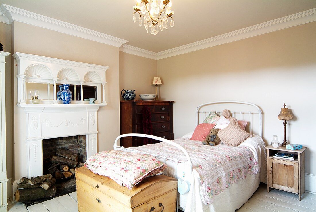 Feminines Schlafzimmer im traditionellen englischen Stil mit offenem elegantem weißem Kamin, antiker Schubladenkommode und naturbelassener Holzkommode vor dem weißen Metallbett