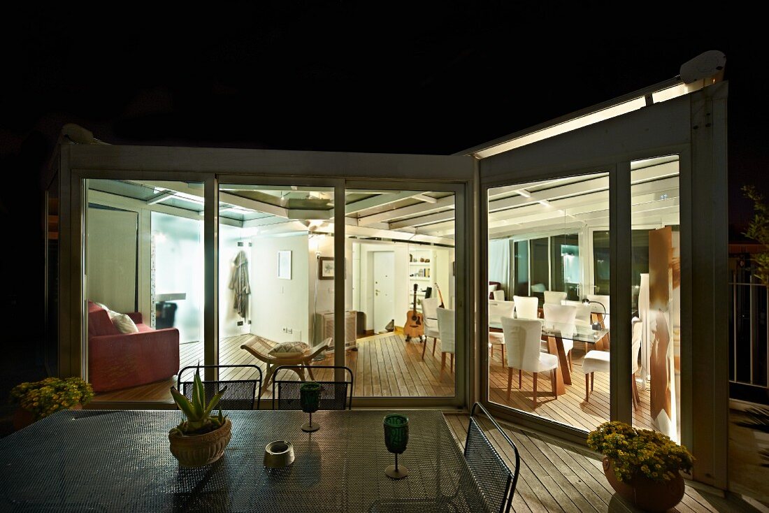 Nächtlicher Blick von der Terrasse mit Gartentisch in den beleuchteten Wohnbereich eines Appartements