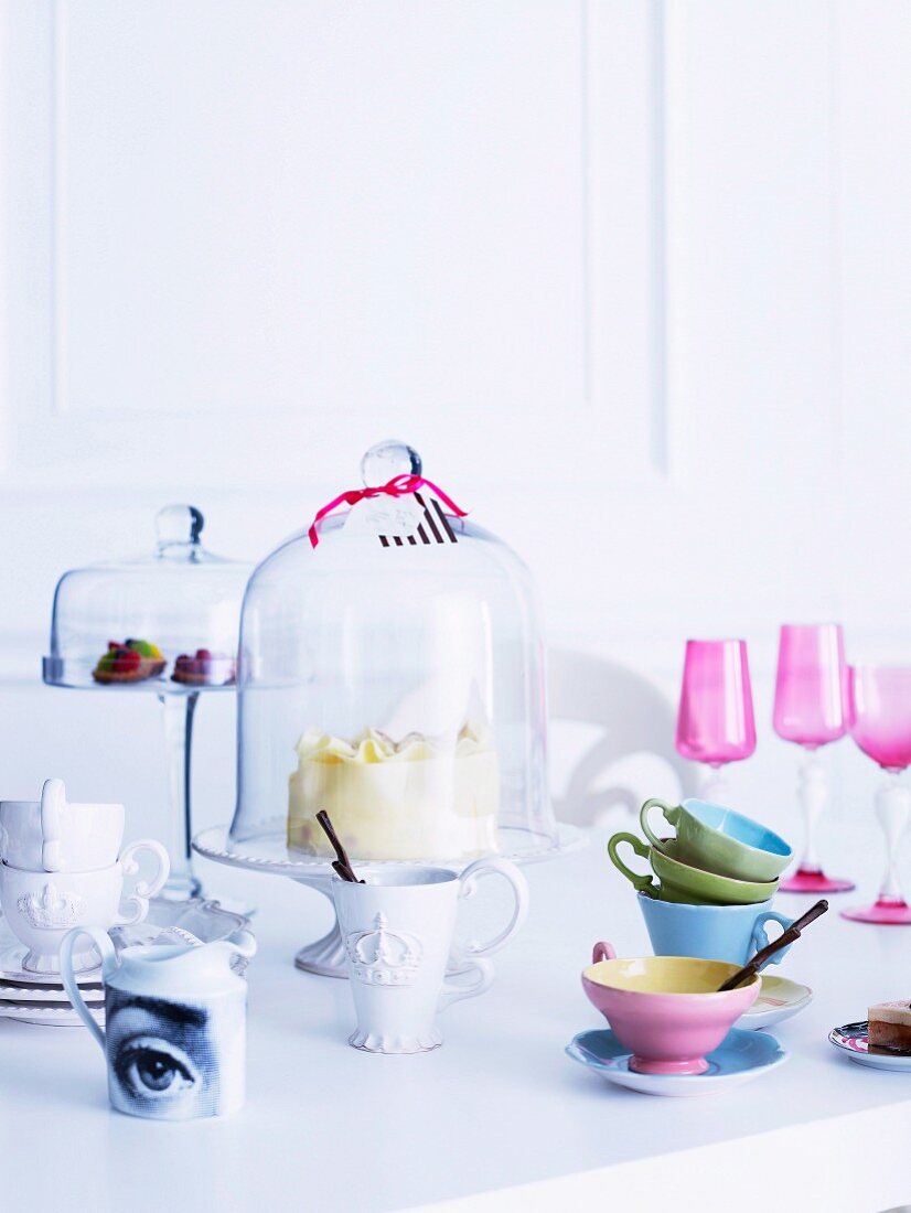 Pastellfarbenes und antikisierendes weisses Porzellan, Etageren mit Glashauben und pinkfarbene Aperitivgläser und bunte Tassen auf einem weißen Tisch