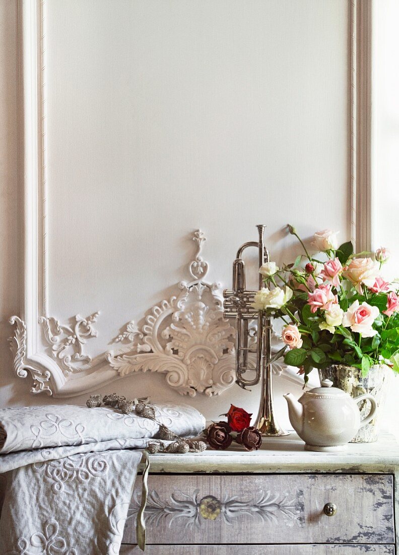 Rosenstrauss und bestickter heller Stoff auf dezent bemalter Kommode vor weißem Holzpaneel mit filigranen Ornamentschnitzereien im eleganten Landhausstil.