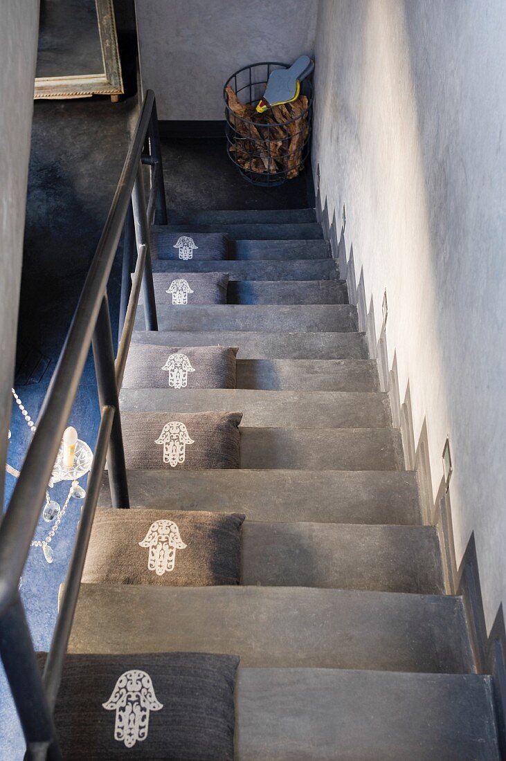 Graue Zierkissen mit aufgedrucktem, glücksbringenden Talisman auf minimalistischer, grauer Treppe mit Metallgeländer