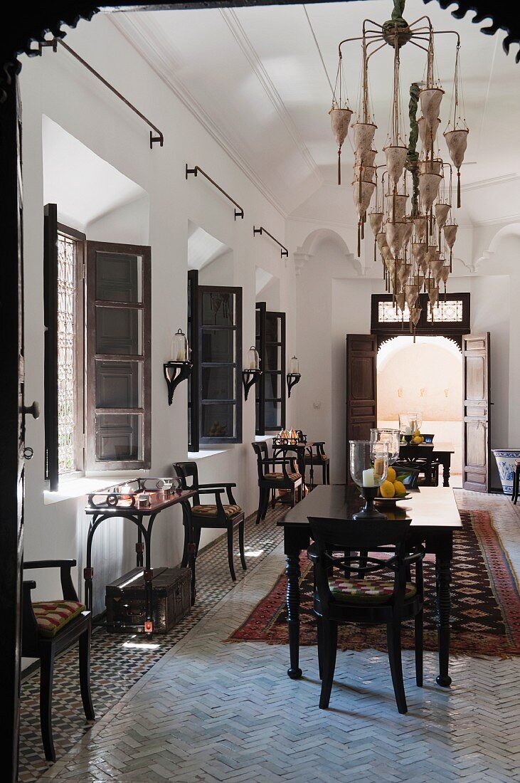 Langezogener Aufenthaltsbereich mit mehreren Fenstern und einer Stuhlreihe im orientalischen Stil mit elegantem dunklem Mobiliar und imposantem Leuchter; Durchblick in den erhellten Innenhof durch die geöffneten Flügeltüren