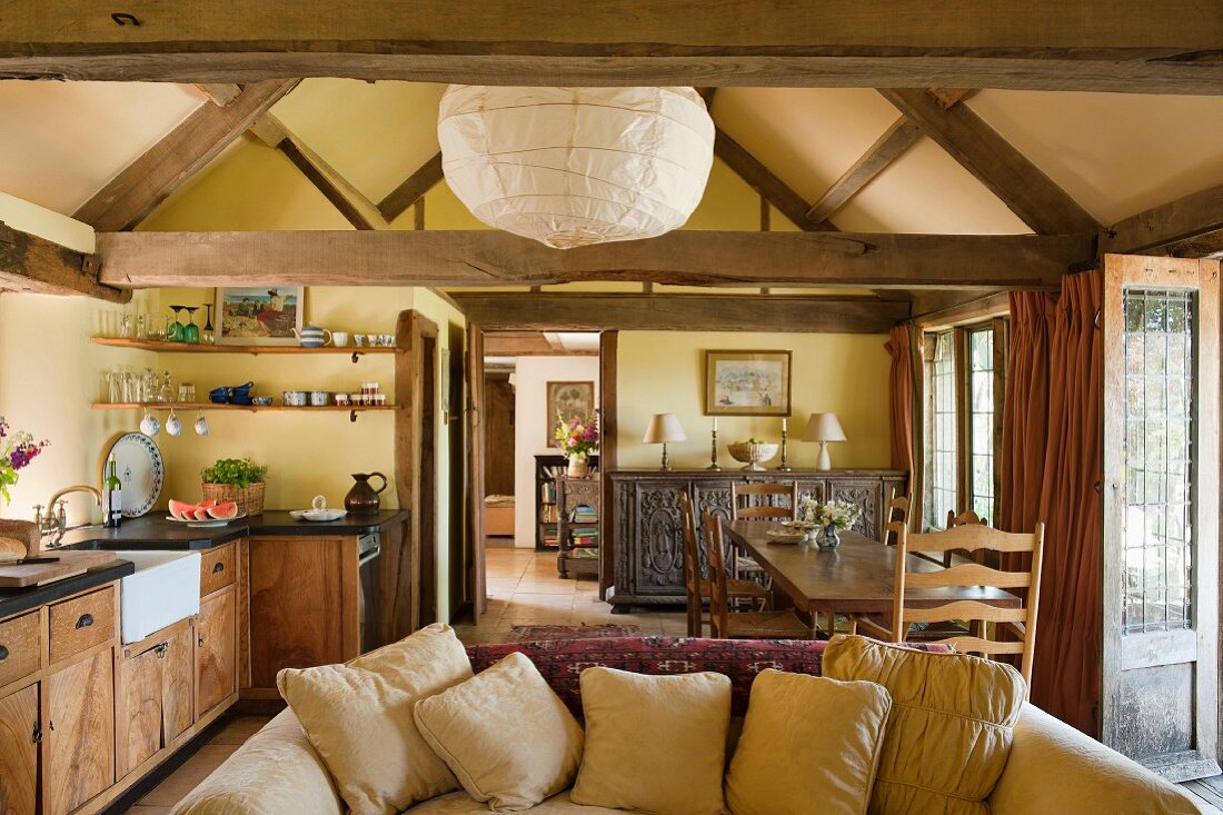 View from kitchen-dining area into foyer of simple English country house with exposed timber structure; comfortable sofa with many scatter cushions in foreground