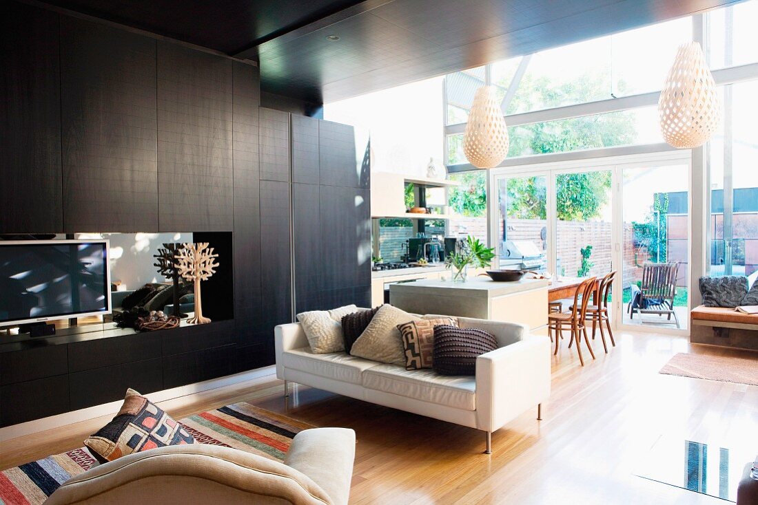 Pale, modern sofa next to black living room cupboards and view into open-plan kitchen-dining room in front of glass facade