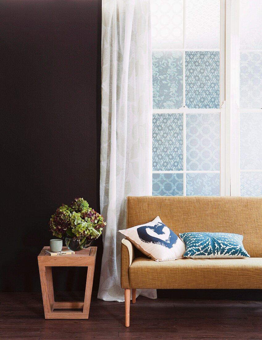 Moderner Beistelltisch aus Holz mit Blumenstrauss neben Sofa im Fiftiesstil vor Fenster mit luftigem Vorhang
