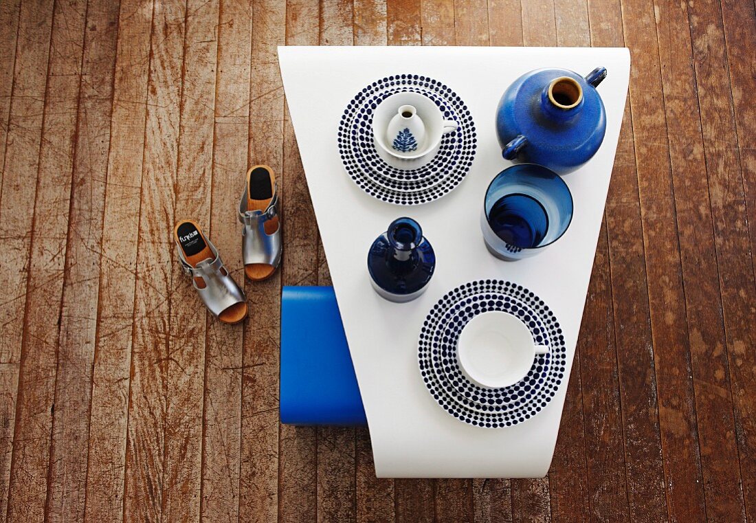 Trapezförmiger, weisser Tisch mit Vintage Geschirr und blauen Vasen auf altem Holzdielenboden; daneben silberne Sandalen