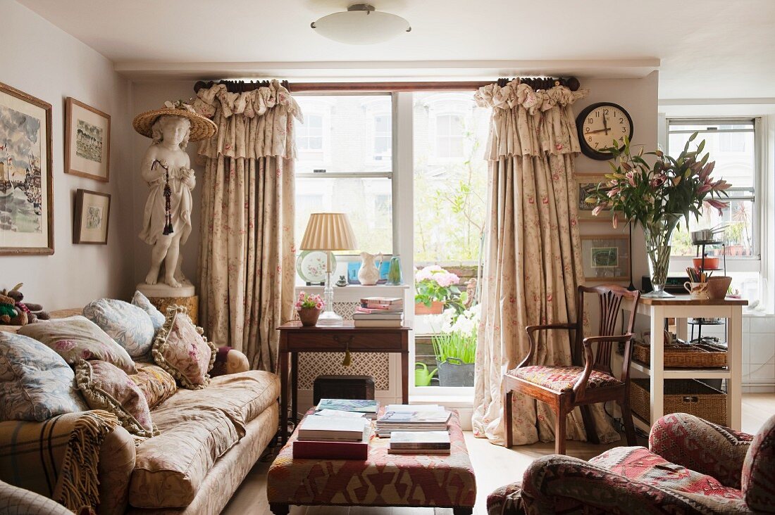 Englisches Wohnzimmer mit Blick auf den Balkon; üppige Volantvorhänge, viele Kissen auf dem Polstersofa und eine Statue in Mädchengestalt, vermitteln ein nostalgisches Flair