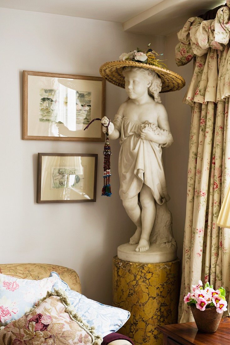 Straw hat on head of statue of girl on pedestal in English living room between valance curtains and sofa with many scatter cushions