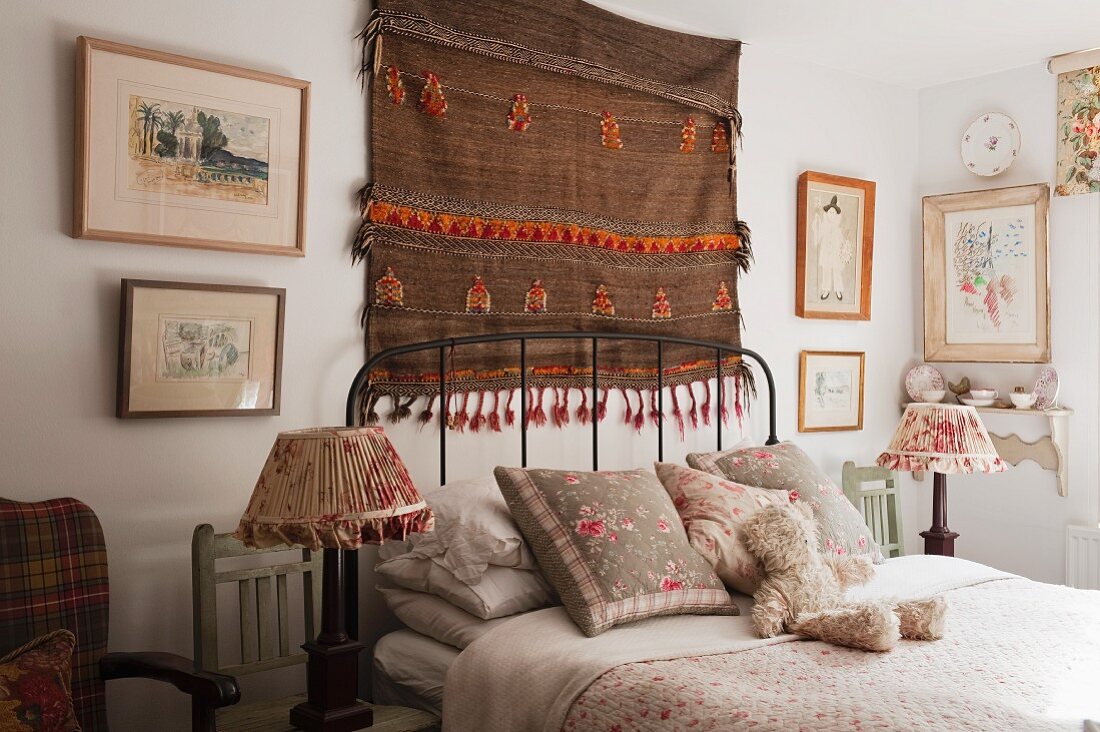 Gewebter überwiegend brauner Wandteppich und gerahmte Bilder über Kopfteil eines gemütlichen Bettes, beidseitig davon nostalgische Nachtischlampen auf Holzstühlen