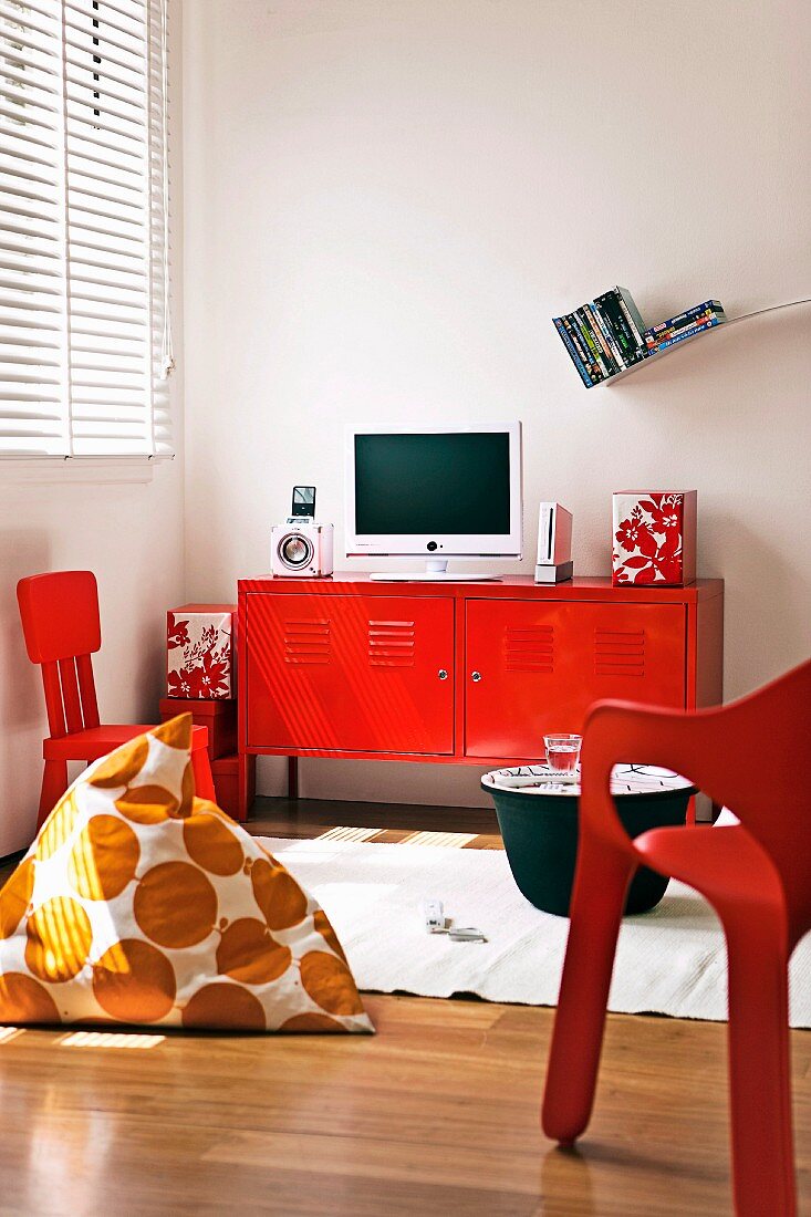 Wohnbereichsecke mit rot lackiertem Metallsideboard und weißem Flachbildschirm; davor rote Kunststoffstühle und ein Aufbewahrungsbeistelltisch mit Tablettcharakter, auf dem glänzenden Parkettboden