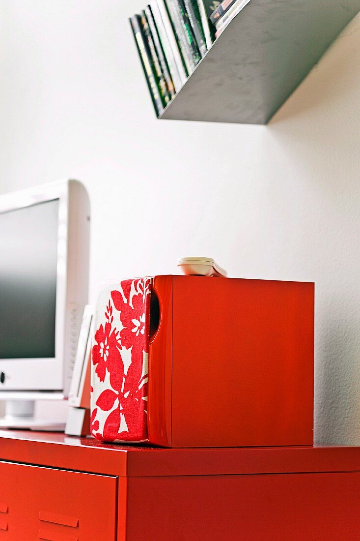 Rotlackiertes Sideboard mit weißem Flachbildschirm und floralgemusterter Box; darüber schräges Metallwandboard mit DVDs