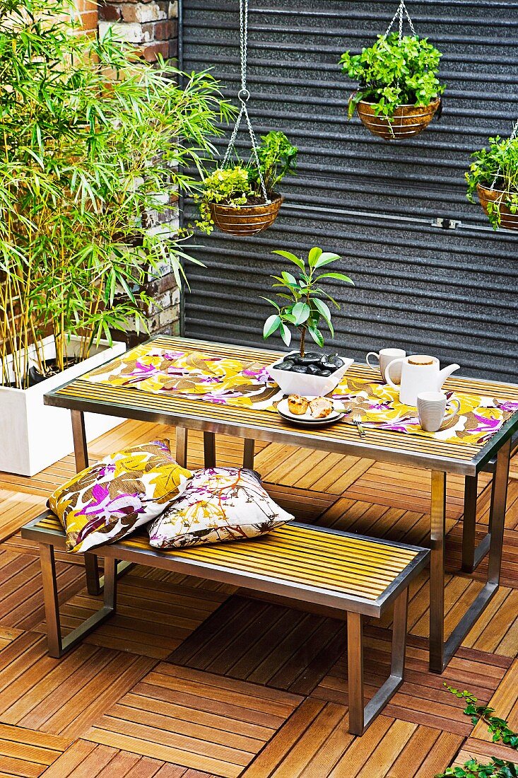 Tiled patio with dining table and benches; in the background a white box with bamboo plants and hanging plant pots