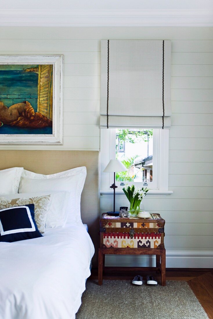 Schlafzimmer mit schmalem Fenster und halb heruntergelassenem Rollo neben Doppelbett mit gepolstertem Kopfende; daneben ein dekoratives Nachtkästchen
