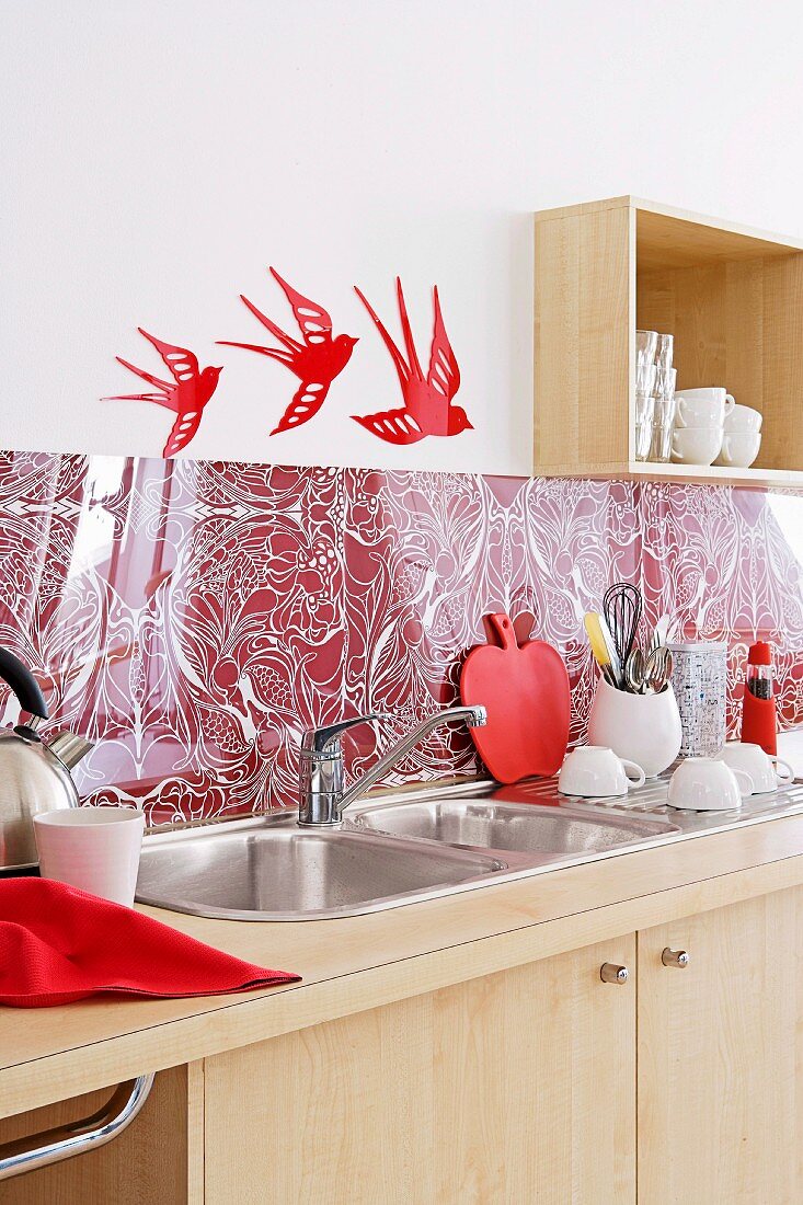 Psychedelische Muster auf rotweissem Spritzschutz über einer Küchenspüle mit ausgeschnittenen Schwalbenmotiven darüber an der weißen Wand.
