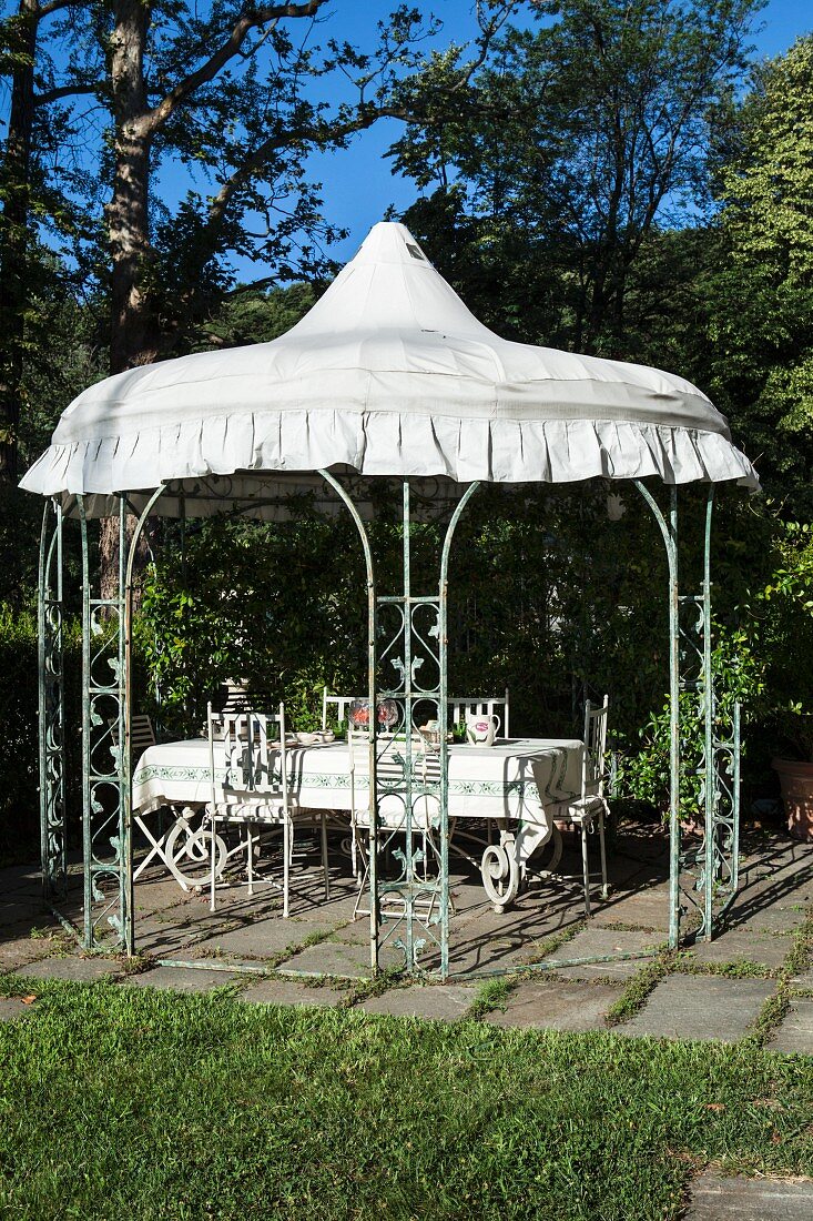 Bewachsener Gartenpavillon mit weißem Dach und darunter gedecktem Esstisch mit Stühlen im Sonnenlicht