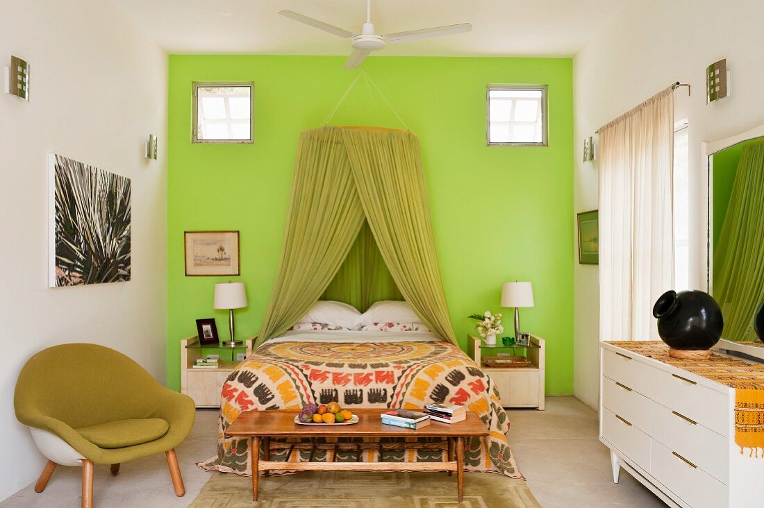 Bedroom with green mosquito net over double bed against green wall and flanked by green shell chair and white sideboard