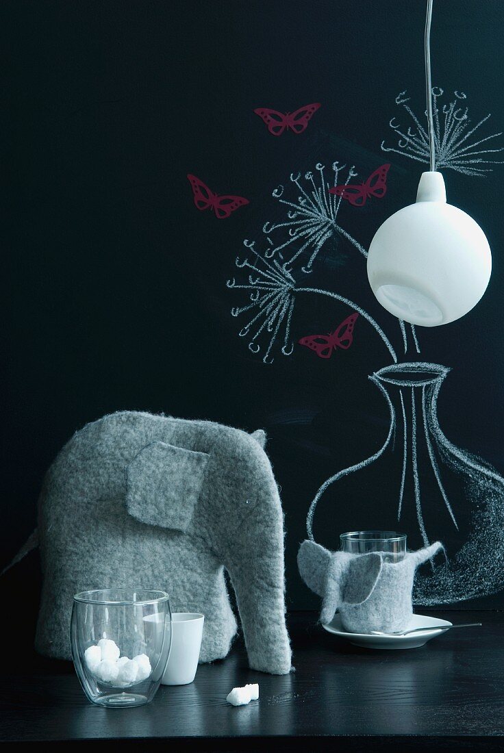 Felt elephant, glasses and minimalist pendant lamp in front of blackboard with floral drawing