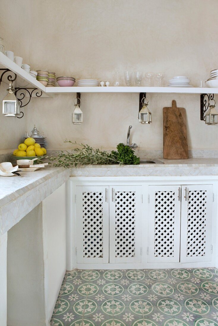 Marokkanische Küchenecke mit heller gefliester Arbeitsplatte und weissen Unterschränken aus Lochholzplatten und floral gemustertem grau-weißem Steinboden; geschwungene Wandkonsolen halten das umlaufende Geschirrboard