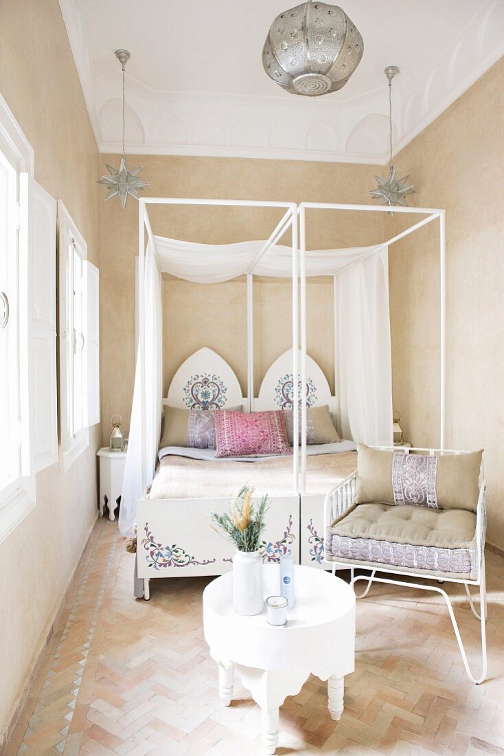 Orientalische Himmelbetten mit bemaltem Kopfteil in hellem, schlichtem Schlafzimmer mit Steinmosaikboden und gepolstertem Metallstuhl neben weißem Beistelltischchen