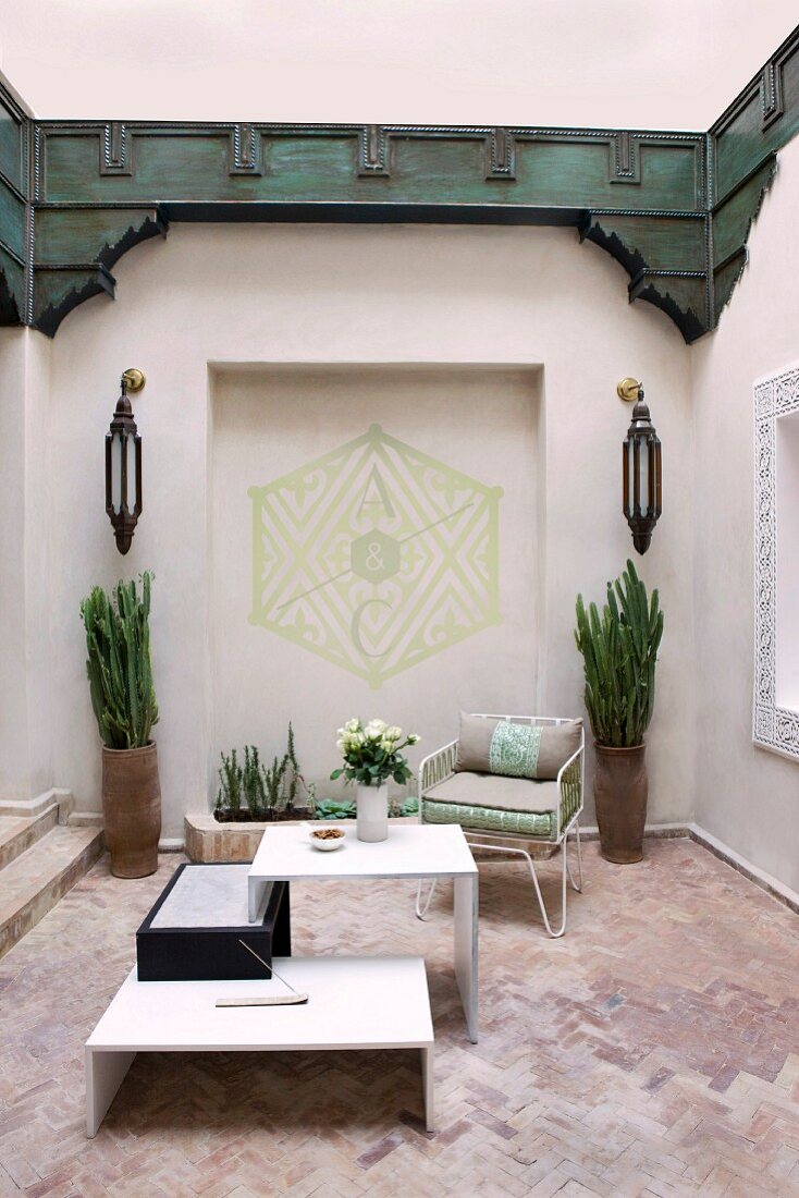 Orientalischer Innenhof mit weißem Designer Tischmöbel auf Terracottaboden; Wandnische mit geometrischer Bemalung und symmetrischen Wandleuchten und Pflanzvasen, sowie umlaufender dunkelgrüner Holzverkleidung an den Wänden