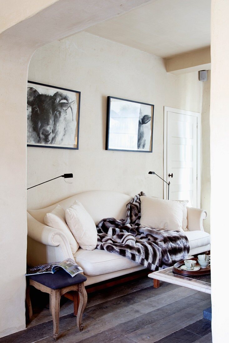 Elegant white sofa and antique, upholstered stool beneath two black and white photographs