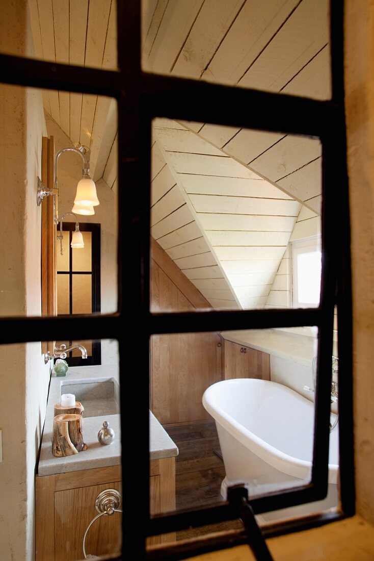 View through an old iron window into the elegant yet cozy bathroom of a country home with a wood panel, pitched ceiling