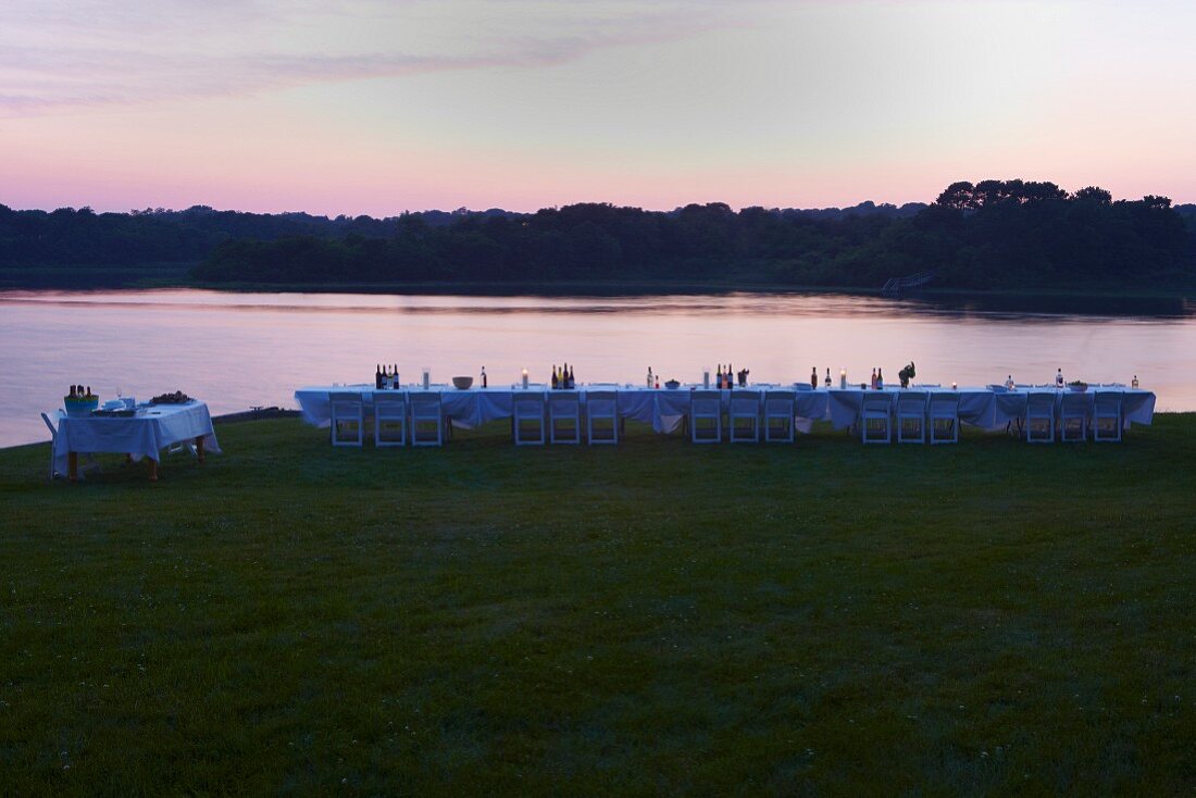Lange Festtafel mit weissen Klappstühlen am Meer bei Sonnenuntergang