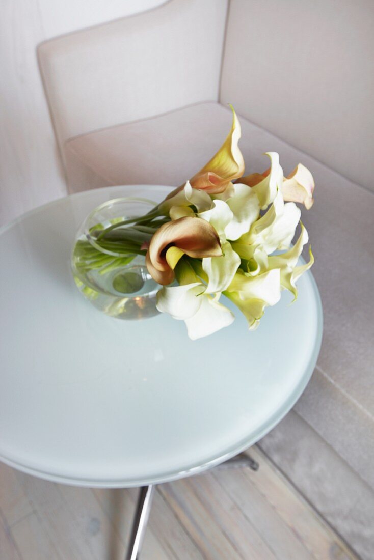 weiße und cremefarbene Calla-Blumen in einer Kugelvase auf einem Milchglastisch